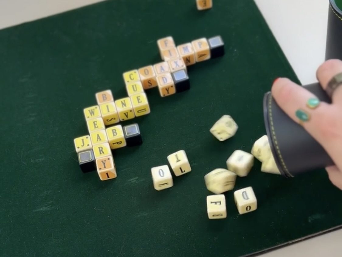 A hand mid-roll, dice tumbling out of a cup onto a green tabletop where a crossword of yellow letter tiles is already laid out